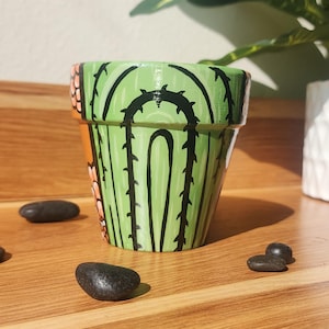 May include: Hand-painted ceramic plant pot with a cactus design. The pot features a green cactus with black spines and orange flowers. The pot is sitting on a wooden surface with black stones.