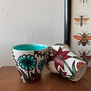 Pode incluir: Dois vasos de flores de terracota pintados à mão com fundo branco e desenhos florais coloridos. Um vaso apresenta flores vermelhas e o outro apresenta flores turquesa.