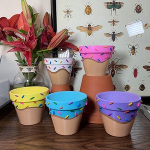 Puede incluir: Cuatro pequeñas macetas de terracota pintadas para parecer conos de helado. Las macetas están decoradas con coloridos espolvoreos y un remolino de glaseado marrón. Las macetas están dispuestas sobre una superficie de madera.