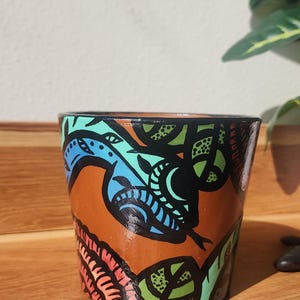 Puede incluir: Maceta de terracota pintada a mano con diseño de serpiente. La maceta tiene una base marrón y una ilustración detallada de serpiente en azul, verde y negro. El diseño incluye detalles en rojo y naranja. Ideal para plantas.