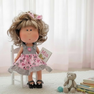 May include: A doll with curly brown hair and a pink bow, wearing a dress with a black and white striped bodice and a pink skirt featuring a heart pattern. The doll holds a small book labeled Mia and wears black shoes while sitting on a white chair.