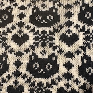 Puede incluir: Primer plano de una tela de punto negra y crema. El patrón presenta caras de gato y formas de corazón que se repiten. El diseño es un patrón repetitivo de caras de gato negras y corazones sobre un fondo crema.
