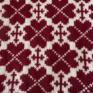 Könnte beinhalten: Ein gestrickter Stoff mit einem sich wiederholenden Muster aus dunkelroten Herzen und weißen kreuzartigen Formen. Das Textil weist ein dichtes, strukturiertes Gewebe mit einem durchgehenden geometrischen Design und einem starken Farbkontrast auf.