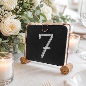 May include: A square black chalkboard table number sign featuring the white chalk-drawn number 7. The board is suspended by a thin copper-colored metal frame, which is supported at the base by two cylindrical cork stoppers on each side.