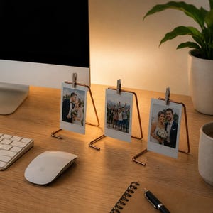 May include: Three copper-colored photo holders displaying instant photos on a wooden desk. A computer, keyboard, and mouse are visible. A potted plant and a mug of coffee are also on the desk. The photos appear to be of a wedding and a group of people.