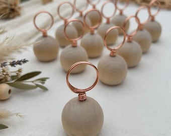 May include: A collection of place card holders, each featuring a smooth, light-colored wooden sphere base topped with a coiled copper-colored wire loop designed to securely hold a small card or paper item in an upright position.