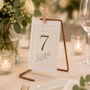 May include: A rectangular white table number card with the text MESA 7 and a green leaf illustration, held by a small wooden clothespin on a copper-colored metal wire stand. The card features a clean design with a thin border and dark green font.