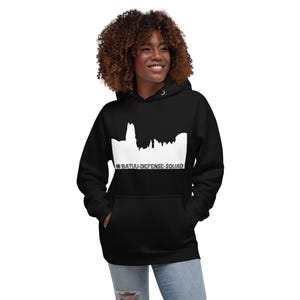 May include: Black hooded sweatshirt with a white graphic of a skyline and the text "#BATUU-DEFENSE-SQUAD". The sweatshirt has a front pocket and a drawstring hood. The model is wearing blue jeans.