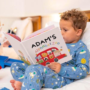 May include: A hardcover children's book titled Adam's Alphabet Zoo featuring a red bus illustration filled with various animals on the cover. The book is held open, displaying colorful graphics of a bus, a zebra, a lion, and a tiger.