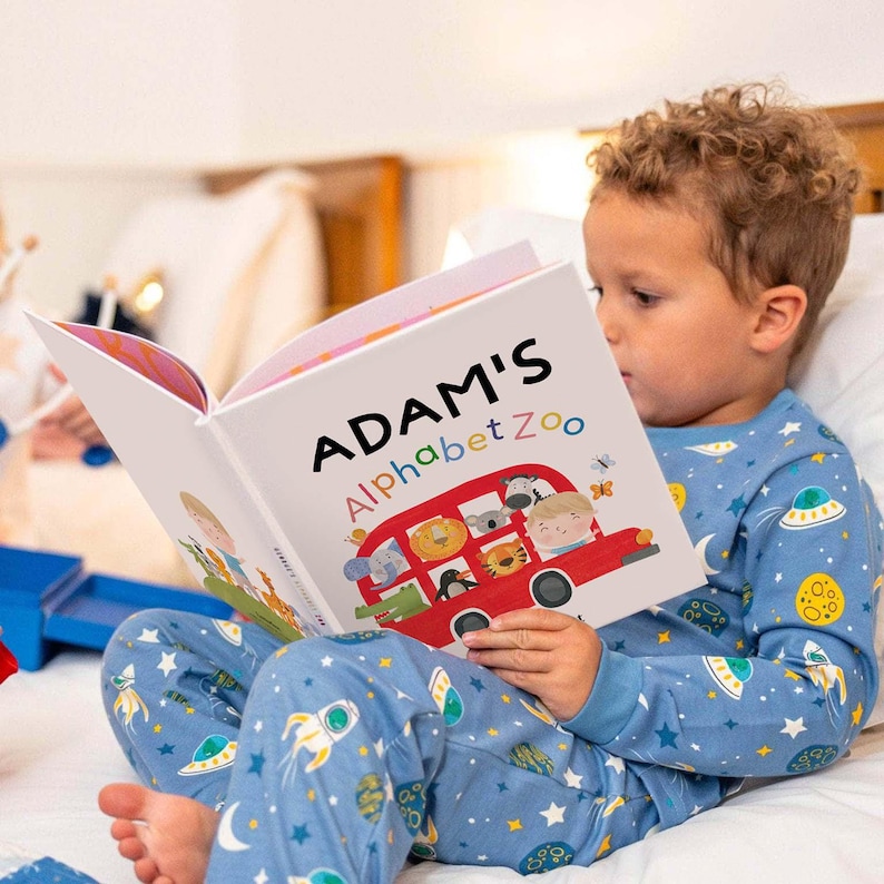 May include: A hardcover children's book titled Adam's Alphabet Zoo featuring a red bus illustration filled with various animals on the cover. The book is held open, displaying colorful graphics of a bus, a zebra, a lion, and a tiger.