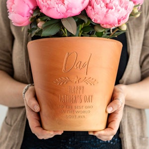 May include: A terracotta flower pot with the word "Dad" engraved at the top, and "Happy Father's Day" below. The pot is filled with pink peonies. The inscription also includes "To the best dad in the world, Love Ava."