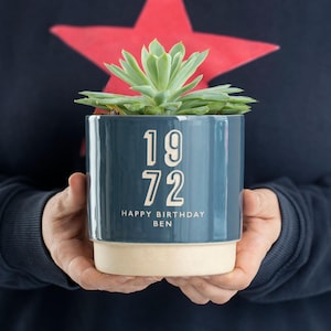 May include: A blue ceramic planter with a white rim, featuring the text "1972 Happy Birthday Ben" in a cream color. The planter is filled with a green succulent plant.