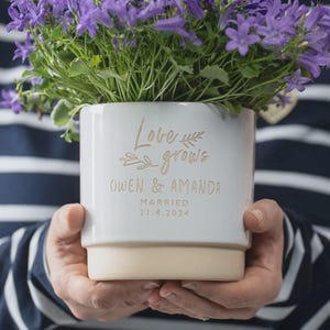 May include: A white ceramic plant pot with a tan base, containing purple bellflowers. The front displays gold text reading Love grows, OWEN & AMANDA, MARRIED, 21.4.2024, with small leaf illustrations. The object is a simple, cylindrical container.