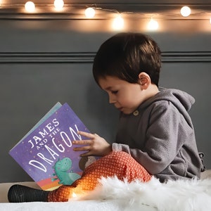 May include: A young child sits on a white fluffy rug, reading a purple book titled "James and the Dragon". The book features a colorful illustration of a green dragon with a rainbow background. The child is wearing a gray hooded sweatshirt and red pants with a white floral pattern.