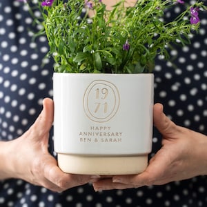 May include: A white ceramic plant pot with a beige base, containing a green plant with purple flowers. The pot is inscribed with "1971" inside a circle, and the words "HAPPY ANNIVERSARY BEN & SARAH".