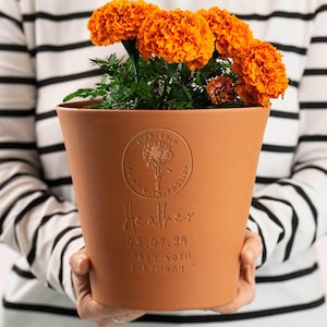 May include: A terracotta flower pot containing bright orange marigold flowers. The pot features an embossed circular logo with a flower illustration, the name Heather, the date 03.07.84, and the text HAPPY 40TH BIRTHDAY engraved on the front surface.