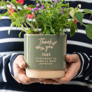 May include: A green ceramic planter with a white rim and the text "Thank you Anna Godparent to Isabella Rose Johnson" in pink lettering. The planter is filled with green plants and pink flowers.