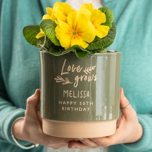 May include: A green ceramic planter with the text "Love grows Melissa Happy 50th Birthday" and a yellow primrose plant inside.
