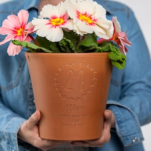 May include: A brown terracotta plant pot with the number "21" in a wreath, and the name "Ellen" engraved below. The pot contains pink and white flowers with yellow centers and green leaves. The text "Happy Birthday Love James & Mum" is also engraved.
