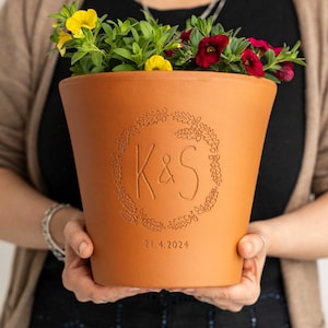 May include: A terracotta plant pot with a personalized design, featuring the initials "K & S" within a wreath and the date "21.4.2024". The pot is filled with vibrant yellow and red flowers and green foliage.