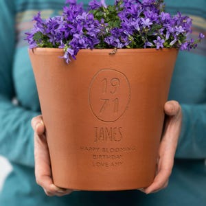 May include: A brown terracotta plant pot overflowing with purple flowers. The pot is inscribed with the year "1971", the name "JAMES", and the message "HAPPY BLOOMING BIRTHDAY LOVE AMY".