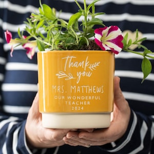 May include: Yellow ceramic planter with white lettering that reads "Thank you, Mrs. Matthews, Our Wonderful Teacher, 2024". The planter is filled with green plants and a pink and white flower.