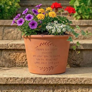 May include: A terracotta flower pot filled with colorful flowers, including purple petunias, yellow marigolds, and red geraniums. The pot has the text "HAPPY BIRTHDAY MUMMY, WE LOVE YOU SO MUCH, YOU'RE AMAZING! LOVE JACK AND LILI" engraved on it.