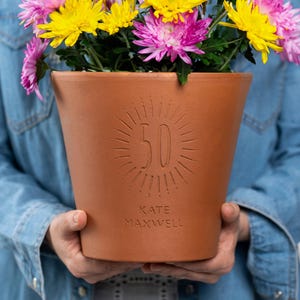 May include: A brown terracotta flower pot with the number 50 and the name "KATE MAXWELL" etched into the front. The pot is filled with yellow and pink chrysanthemums. The pot is held by a person wearing a denim shirt.