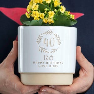 May include: White ceramic planter with a cream-colored base. The planter has a wreath design with the number 40 in the center. The text "Izzy Happy Birthday Love Ruby" is printed below the wreath.