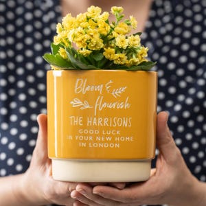 May include: A yellow ceramic planter with the text "Bloom & Flourish The Harrisons Good Luck in your new home in London" in white lettering. A small yellow flowering plant is in the planter.