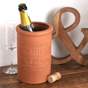 May include: Terracotta wine cooler with the text "The wedding of Isabella and Andrew June 18th 2016" engraved on the side. A bottle of champagne is in the cooler and a cork is on the table.