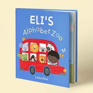 May include: A blue hardcover children's book titled Eli's Alphabet Zoo. The cover features a red bus filled with illustrated animals including an elephant, lion, koala, zebra, crocodile, penguin, and tiger, with a child driving and butterflies.