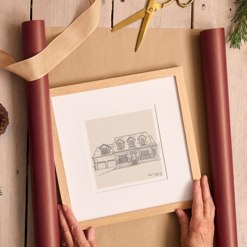 K&ouml;nnte beinhalten: Eine gerahmte Hausskizze mit hellem Holzrahmen, die auf braunem Geschenkpapier liegt. Burgunderfarbene Geschenkpapierrollen und eine goldene Schere sind ebenfalls zu sehen. Die Skizze ist eine detaillierte Strichzeichnung eines Hauses.