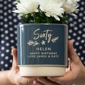 May include: A blue ceramic planter with the text "Sixty * Helen Happy Birthday Love James & Kate" in gold lettering. The planter is filled with white flowers.