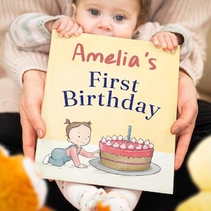 May include: A hardcover book with a pale yellow cover featuring the text Amelia's First Birthday in red and blue lettering. The illustration shows a small child crawling toward a pink frosted cake with a single lit blue candle on a plate.