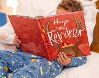 May include: A child in blue pajamas with a rocket and spaceship pattern reads a book titled "Hugo and the Reindeer." The book cover is red with a reindeer illustration and the title in white script. The child is in bed with a white pillow and a wooden headboard.
