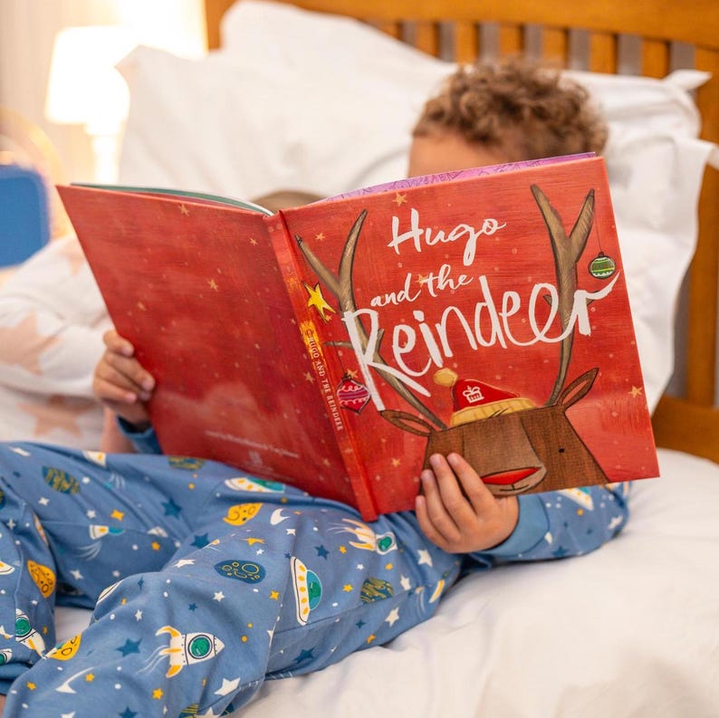May include: A red children's book titled 'Hugo and the Reindeer' features a reindeer illustration wearing a crown. The book cover has a festive design with stars and ornaments.  The book is open, showcasing its colorful pages and illustrations.