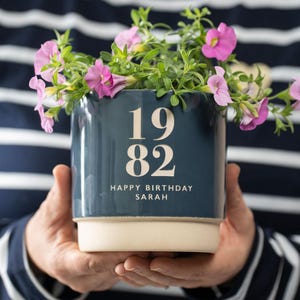 May include: A blue ceramic planter with white lettering that says "1982 Happy Birthday Sarah". The planter is filled with pink flowers.