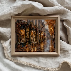 Peut inclure: Une impression d'art encadrée représentant une scène de rue européenne en automne. L'image montre une librairie avec de grandes vitrines, un éclairage intérieur chaleureux et un feuillage orange sur la façade, le long d'une rue pavée humide.