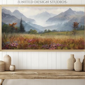May include: A framed landscape painting of a mountain range with a misty valley and wildflowers in the foreground. The painting is in a wooden frame and is hanging on a white wall.