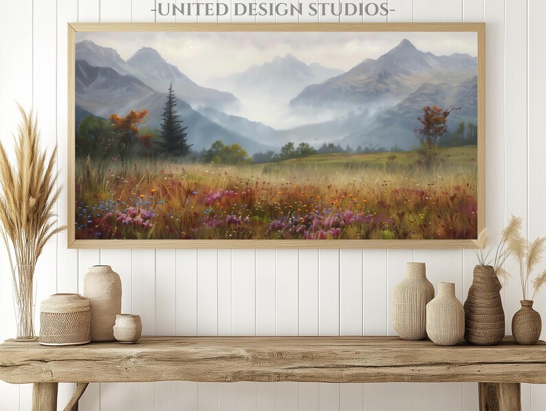 May include: A framed landscape painting of a mountain range with a misty valley and wildflowers in the foreground. The painting is in a wooden frame and is hanging on a white wall.
