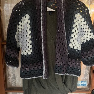 May include: A handmade crochet cardigan in shades of gray, black, and white, with a granny square design. The open-front sweater is embellished with sequins and hangs over a green top. The cardigan is displayed on a hanger.