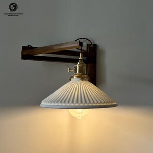 May include: A wall-mounted light fixture with a white, ribbed lampshade and a brass-colored bulb socket. The fixture is attached to a dark brown wooden arm and a rectangular wooden base. The light is on, illuminating the area below.