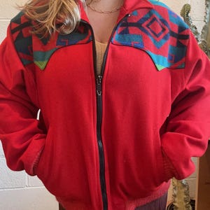 May include: A red zip-up jacket with a black zipper and pockets. The jacket features a geometric pattern in teal, blue, and black on the shoulders. The jacket has a ribbed hem and cuffs.