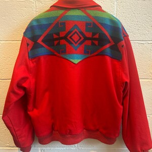 May include: Red jacket with a colorful geometric design on the upper back. The design features a diamond shape in the center, with blue, green, and black accents. The jacket has a ribbed hem and cuffs.