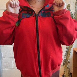 May include: A bright red bomber jacket with a black zipper and a patterned collar. The collar features a design with teal, black, and green accents. The jacket has ribbed cuffs and a ribbed hem.