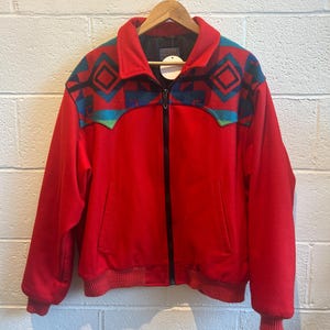 May include: A red zip-up jacket with a geometric pattern in teal, navy, and green on the shoulders. The jacket has a ribbed hem and cuffs, and a black zipper. The jacket is hanging on a wooden hanger.