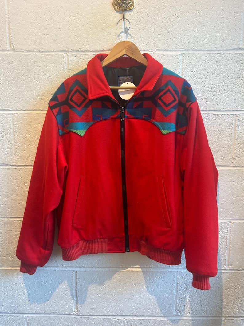 May include: A red zip-up jacket with a geometric pattern in teal, navy, and green on the shoulders. The jacket has a ribbed hem and cuffs, and a black zipper. The jacket is hanging on a wooden hanger.
