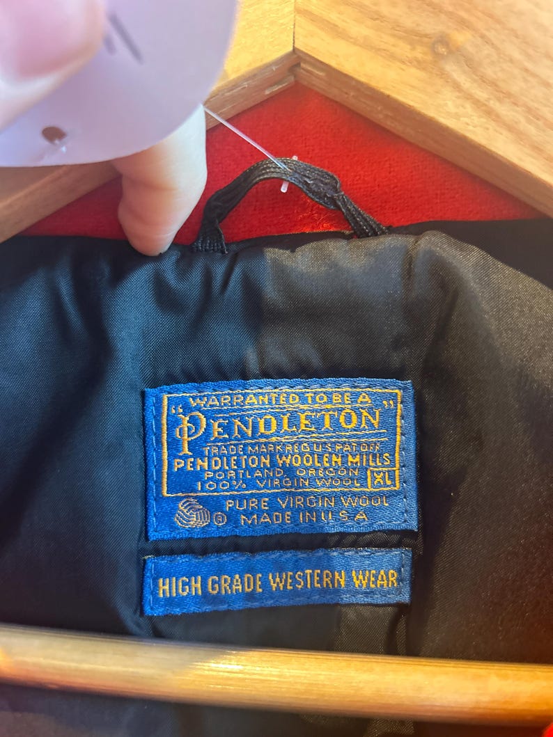 May include: Close-up of a black jacket lining with a blue Pendleton label. The label reads "Warranted to be a Pendleton" and "High Grade Western Wear." The jacket is made of 100% virgin wool and is a size XL.