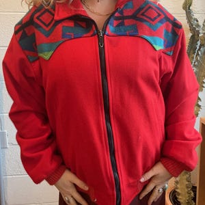 May include: A red zip-up jacket with a geometric pattern in teal, black, and turquoise across the shoulders. The jacket has a black zipper and ribbed cuffs. The jacket is a solid red color.